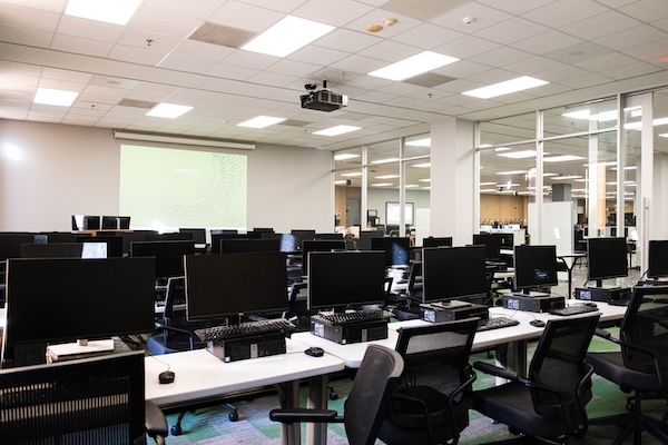 computer classroom with computer stations and presentation screen as seen from rear-left